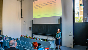 Blick von hinten über die Köpfe der Menschen im Hörsaal nach vorn. Eine weibliche Person steht am Pult um spricht. Über ihr ist eine Folie projiziert mit einem Goethe-Zitat: „Musik im besten Sinne bedarf weniger der Neuheit, ja vielmehr je älter sie ist, je gewohnter man sie ist, desto mehr wirkt sie.“