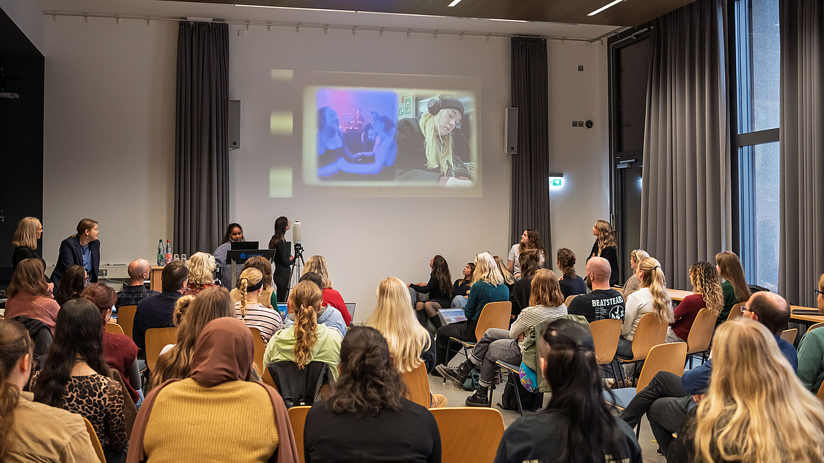 Totale von hinten übder das Publikum hinweg auf dioe Projektiion eines Videos.