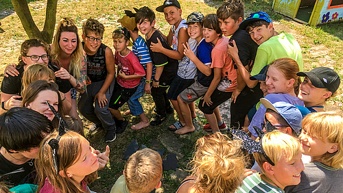 Das Bild zeigt von schräg oben eine Polonaise im Kreis mit 20 fröhlich lachenden Kindern und zwei jungen Frauen. 