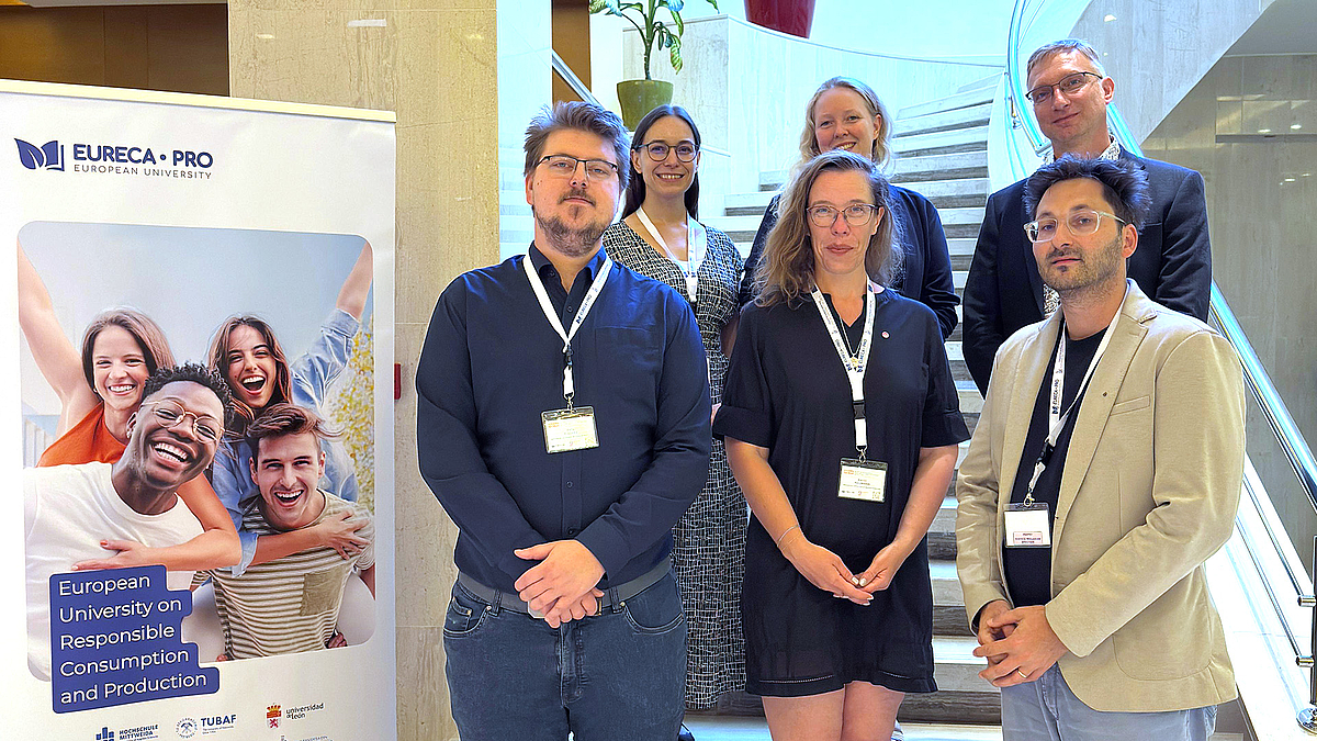 Sechs Menschen stehen auf zwei Stufen einer Treppe und posieren förmlich für ein Gruppenfoto. Links im Bild steht ein Rollup mit einem Bild lachender, fröhlicher Menschen.