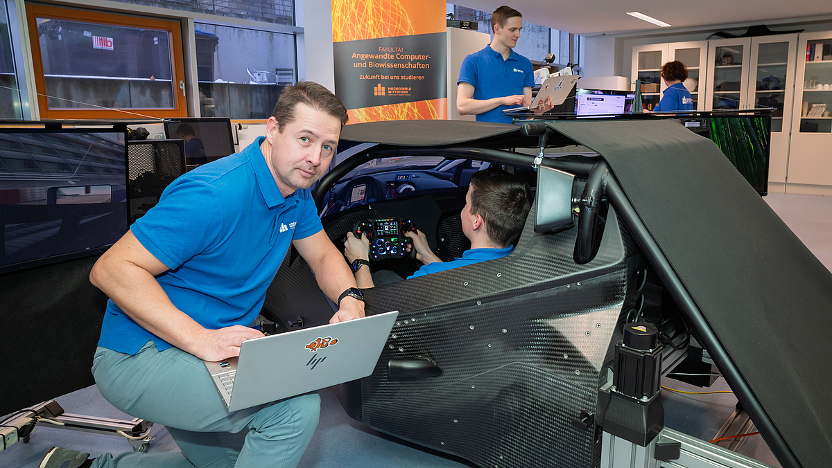 Immersiv: Full-Scale Simulatorplattform eines KTM xBow GT4 im Living Lab der HSMW (Foto: Helmut Hammer) Um einen rennwagenähnlichen Simulator, in dem eine männliche Person sitzt, knien bzw. stehen zwei weitere männliche Personen mit Laptops in den Händen.
