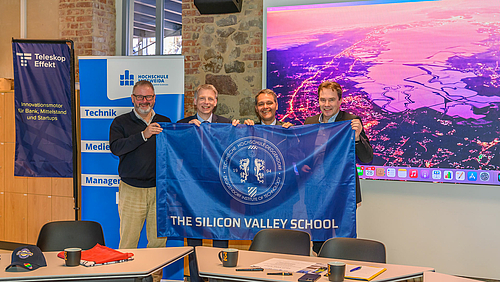 Vier männliche Personen halten freudenstrahlend eine Fahne mit der Ausfschrift "Siliccon Valley School". Im Hintergrund ist eine Luftaufnahme vom Silicon Valley in Klaifornien zu sehen, sowie drzwei Rollups von Teleskopeffekt und Hochschule Mittweida.