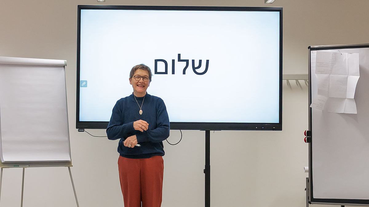 Ein weibliche Person mit kurzen Haaren udn Brille steht vor eiem großen Bildschirm, auf dem  in hebräischer Shrift ausschließlich das Wort Shalom zu sehen ist.