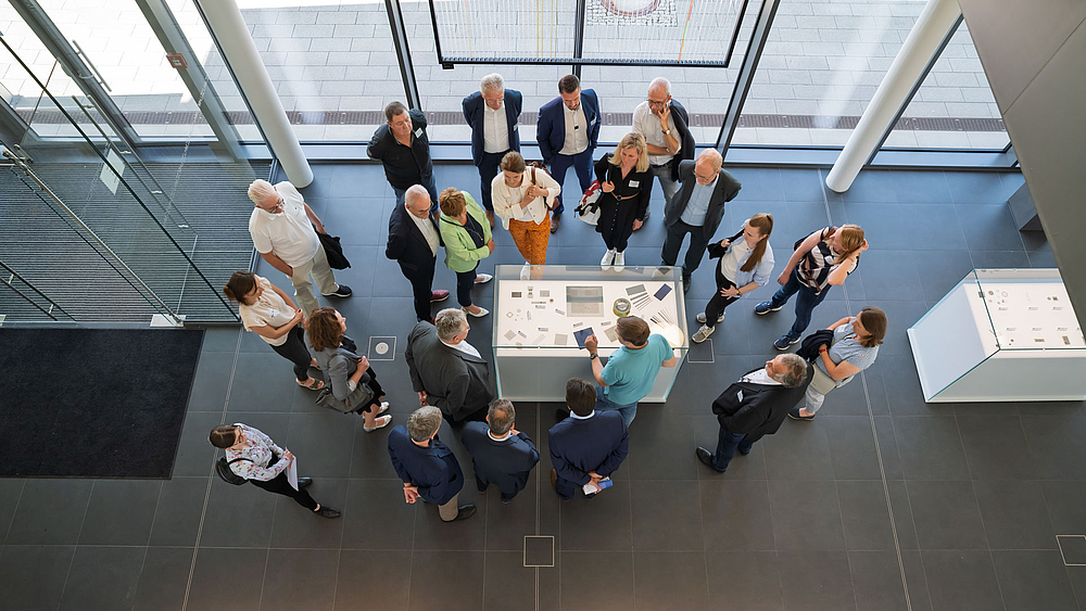 Blick von oben auf eine Gruppe von rund zwanzig Personen, die von oben auf eine Vitrine schauen.