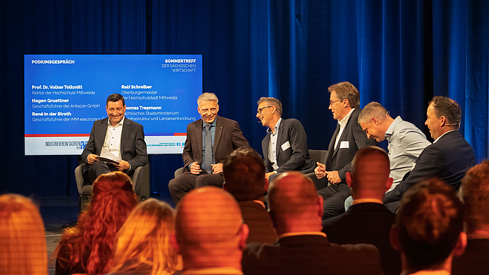 Sechs lachende männliche Personen in Sesseln auf einem Podium in einem Studio. Im Bildvordergrund sind Köpfe von Zuschauenden von hinten zu sehen.