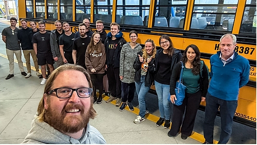 Selfie zeigt eine Gruppe von überwiegend jungen Personen vor einem typischen gelben us-amerikanischen Schulbus. Im Bildvordergrund ist der Kopf des Fotograf zu sehen.
