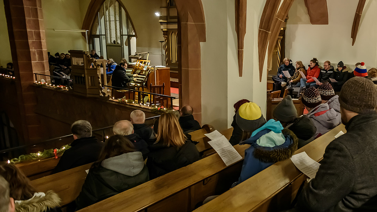 Menschen in Kirchenbänken mit Liedblättern. Im Hintergrund der Organist am Spieltisch.