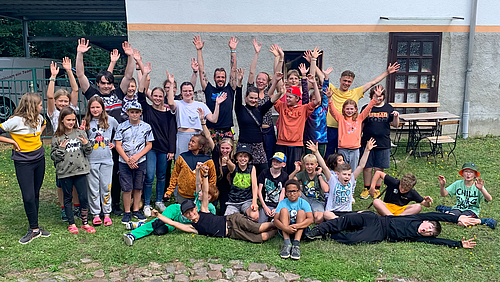 Gruppenbild auf einer Wiese vor einem Gebäude mit rund 30 Personen, darunter überwiegend Kinder zwischen 8 und 12 Jahren. Die meisten stehen jubelnd mit nach oben gestreckten Armen, andere sitzen auf dem Boden, einige liegen. 