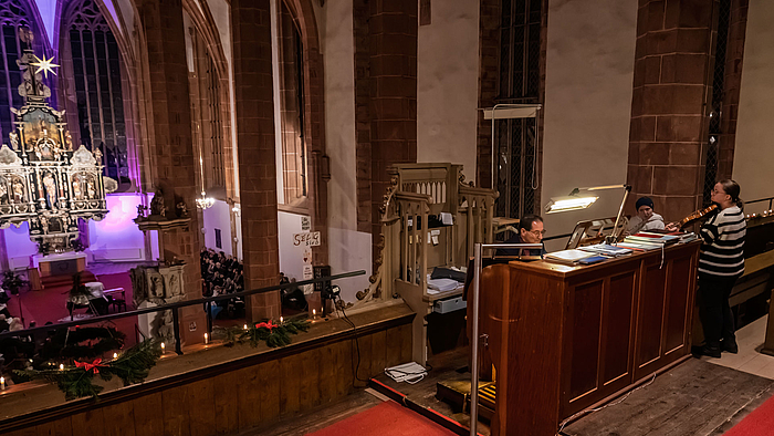 Blick auf Empore und Kirchenschiff. Auf der Empore sitzt eine männliche Person am Spieltisch der Orgel, rechts daneben stehen zwei weibliche Personen, eine spielt Geige, die andere Querflöte.
