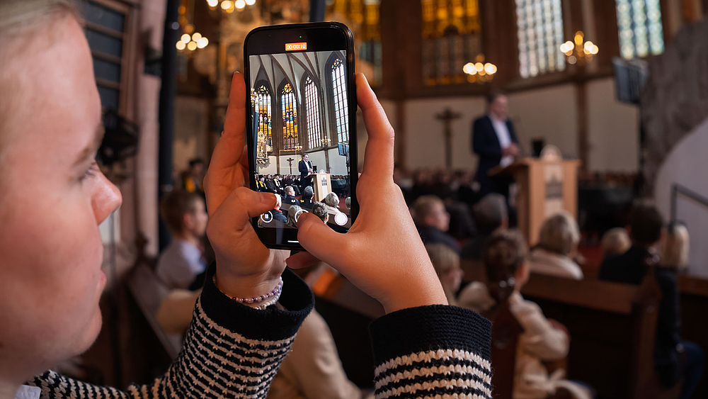 Blick auf den Smartphone-Bildschirm einer weiblichen Person, die einen männlichen Redner im Altarraum filmt.