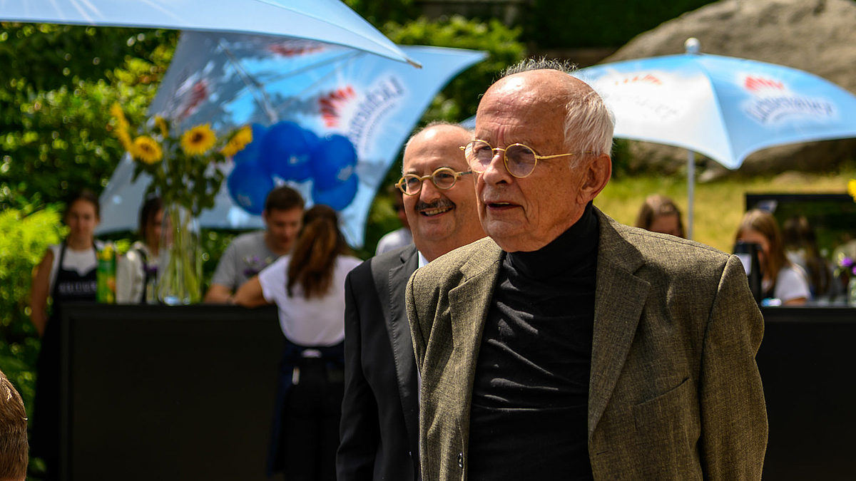 Auf dem Foto stehen zwei männliche Personen leicht versetzt hintereinander, beide in die gleiche Richtung schauend.