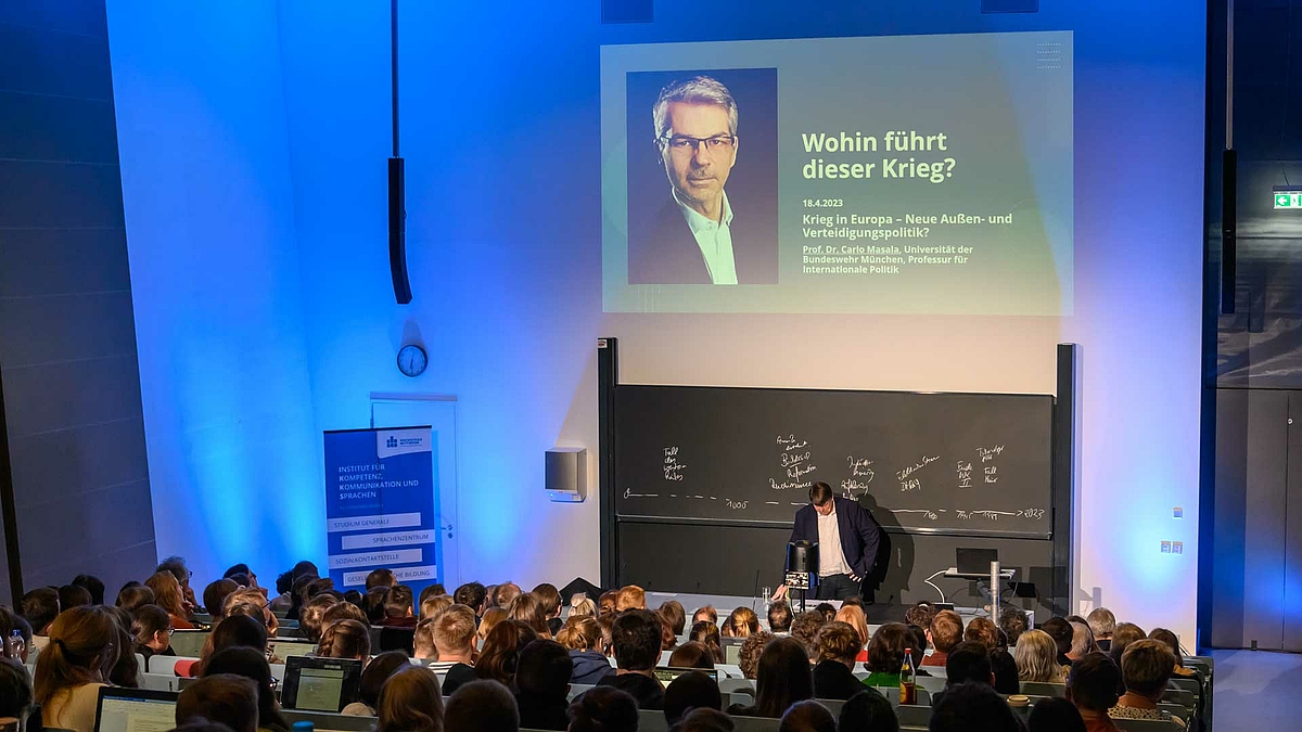 Blick von hinten über die Köpfe der Zuschauenden auf den Dozenten, der vor der Tafel steht. Über ihm zeigt die Projektion links das Portrait einer männlichen Person und rechts die Worte „Wohin führt dieser Krieg? 18.04.2023. Krieg in Europa – Neue Außen- und Sicherheitspolitik? Prof. Dr. Carlo Masala. Universität der Bundeswehr München. Professur für internationale Politik.“