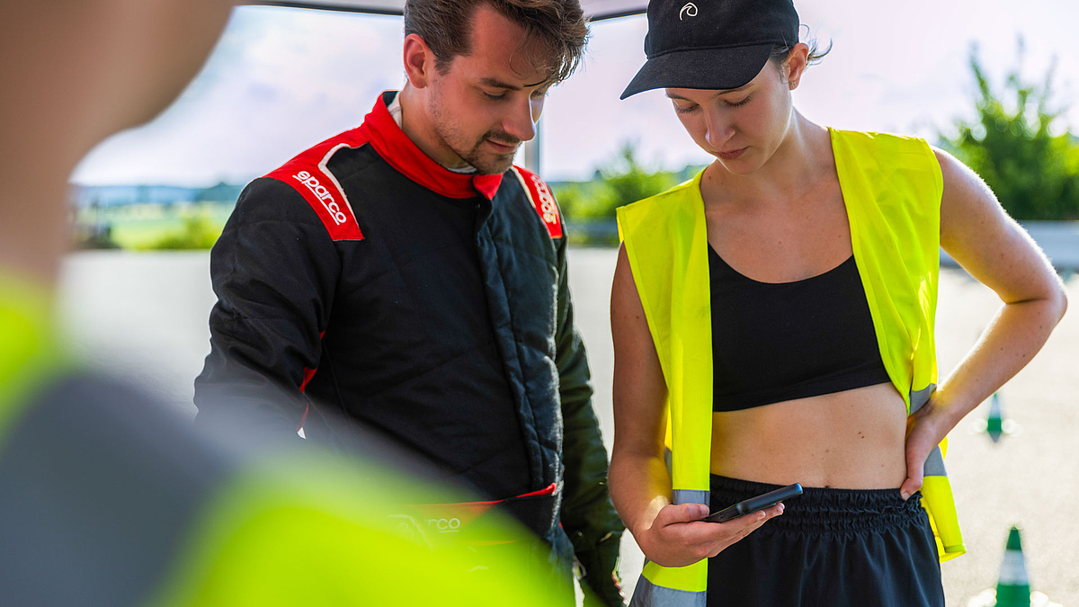 Eine junge männliche Person im Rennoverall und eine junge weibliche Person in Top und neongelber Weste stehen nebeneinander und schauen gemeinsam in ein Smartphone, das die weibliche Person in der Hand hält. Im Hintergrund sind unscharf weiß-rote Pylonen zu sehen.