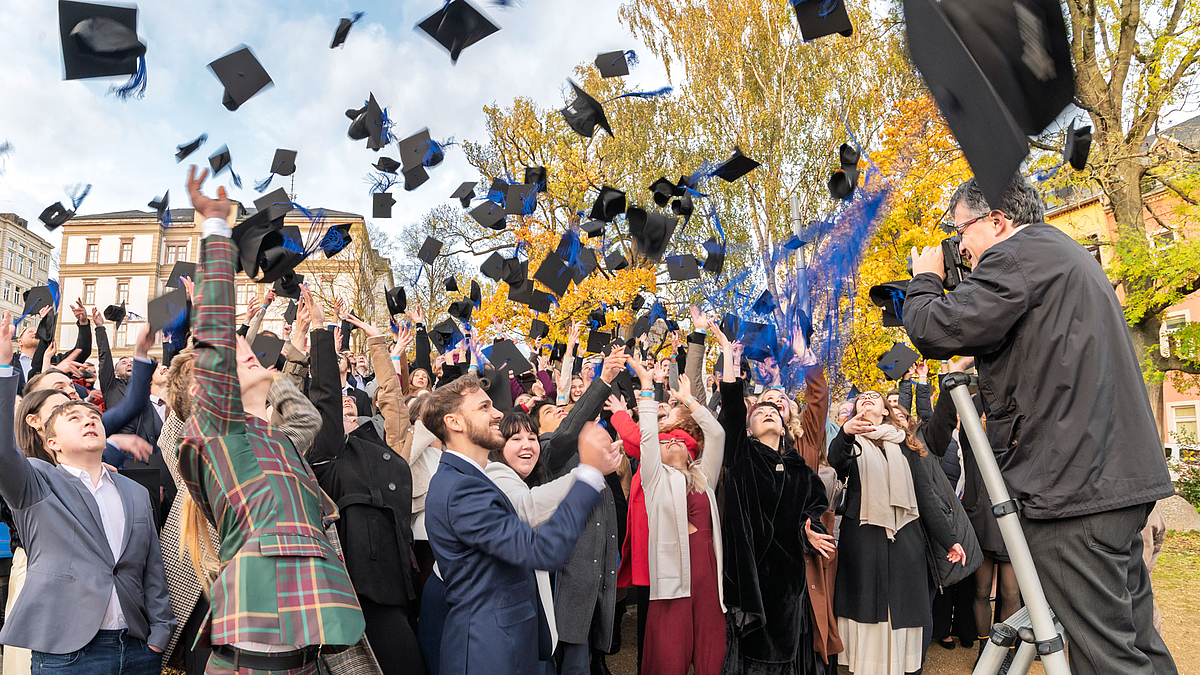 Fotograf auf der Leiter fotografiert die diertkt vor im stehenden Absolvent:innen beim Hochwerfen der Hüte.