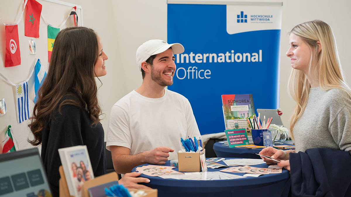 Zwei junge weibliche und eine junge männliche Person an einem Stehtisch unterhalten sich fröhlich.