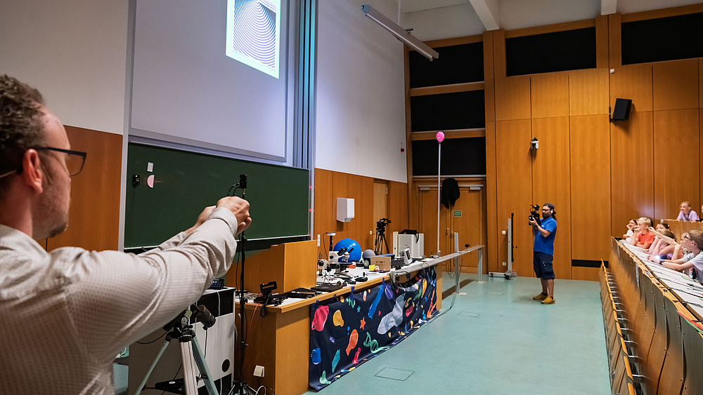 Dozent richtet einen Laserstrahl auf einen Ballon, der an einer Stange befestigt ist. Der Lichtstrahl ist als heller Punkt auf dem Ballon zu erkennen.