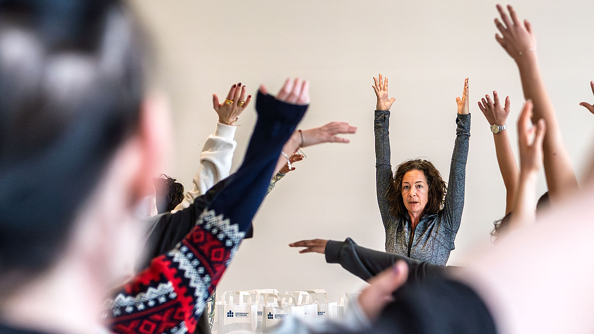 Nach oben getzsrecjte Arme verscheidner Personen von hinten zu segen. Im Bildehintergrund ist die Übungsleiterin zu segen.