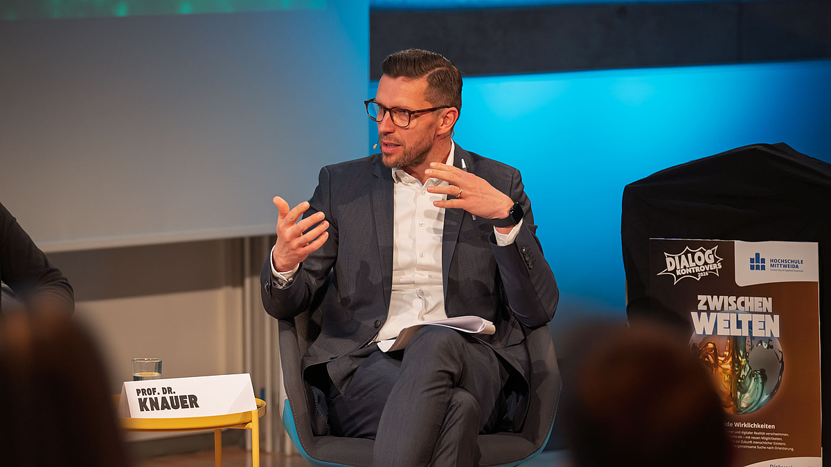 Männliche Person im Podium, gestikulierend, nenne ihr das Plakat von der Veranstaltung mit dem Text "Dialog Kontrovers 2026. Zwichenwelten".