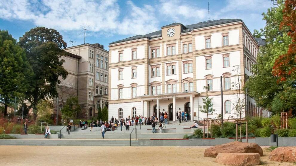 Hochschule Mittweida – Hauptgebäude mit Uhr und Treppenanlage, von Bäumen umgeben bei blauem Himmel