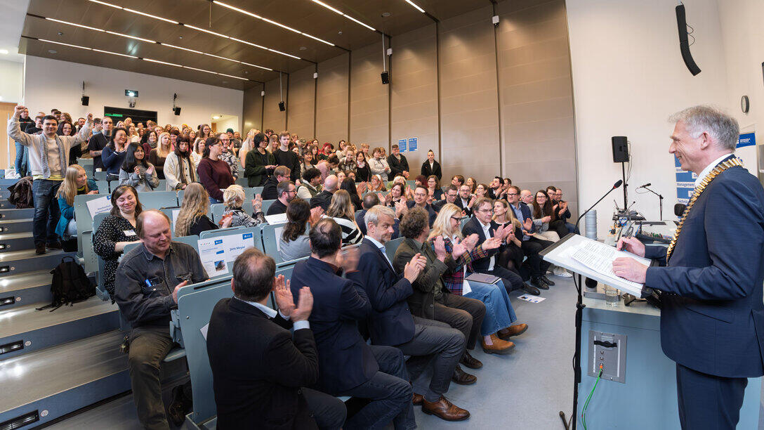 Blick in den Hörsaal mit vielen in den Bankreichen stehenden Menschen. Am unteren Ende des Hörsaals und rechts im Bild eine männliche Person im Anzug und mit goldener Amtskette am Rednerpult.