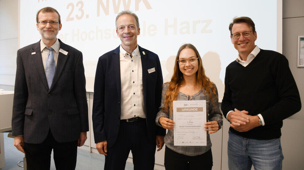 Drei männliche Personen und eine weibliche Person stehen vor einem beleuchteten Hintergrund, die weibliche Person hält eine Urkunde in den Händen