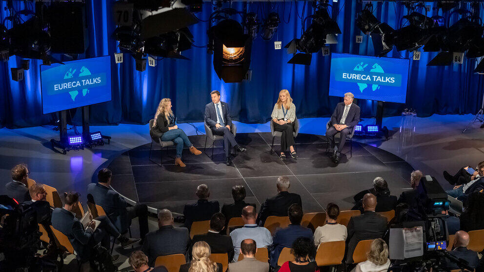 Four people on a podium in a television studio. Spectators can be seen from behind in the foreground.