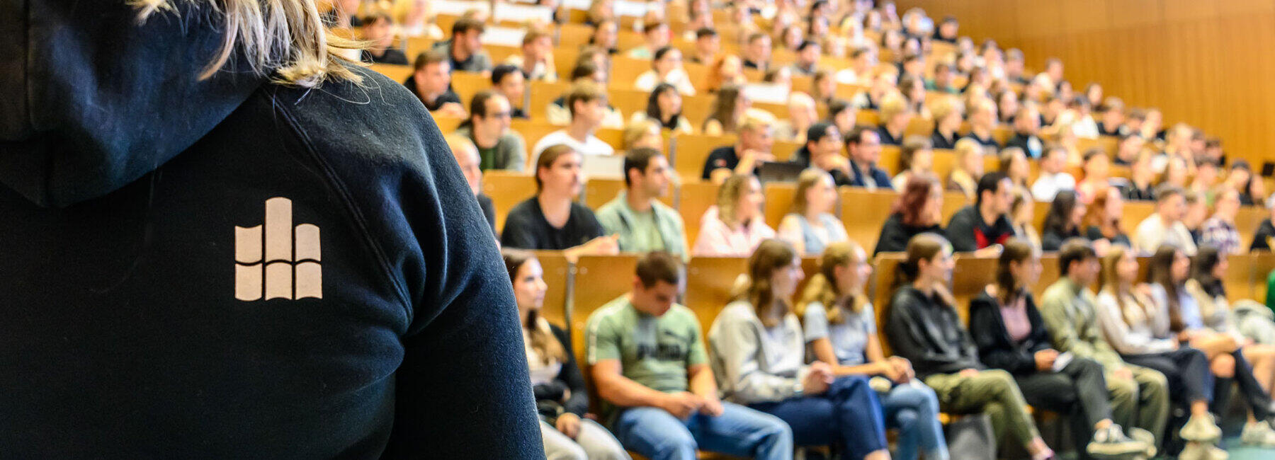 Die Bankreihen eines Hörsaals sind voll besetzt. Am linken Bildrand steht eine Person, die sich den Pesonen im Hörsaal zuwendet. Sie trägt einen blauen Kapuzenpullover mit dem Hochschullogo auf dem Rücken.