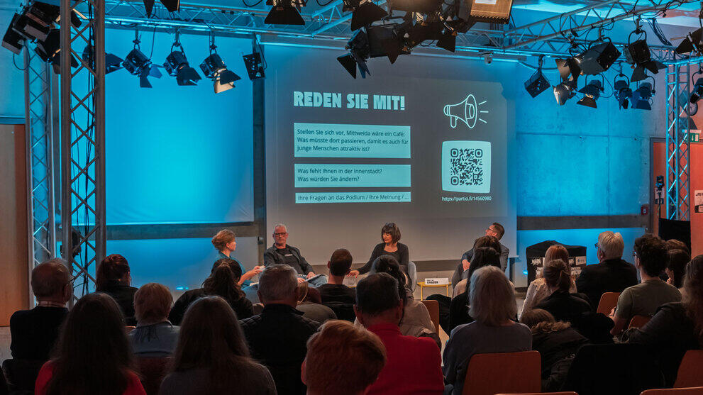 Blick von hinten über die Köpfe des Publikums hinweg auf das Podium. Auf der Leinwand dahinter ist zu lesen "REDEN SIE MIT!", darunter drei Fragen und daneben ein QR-Code.