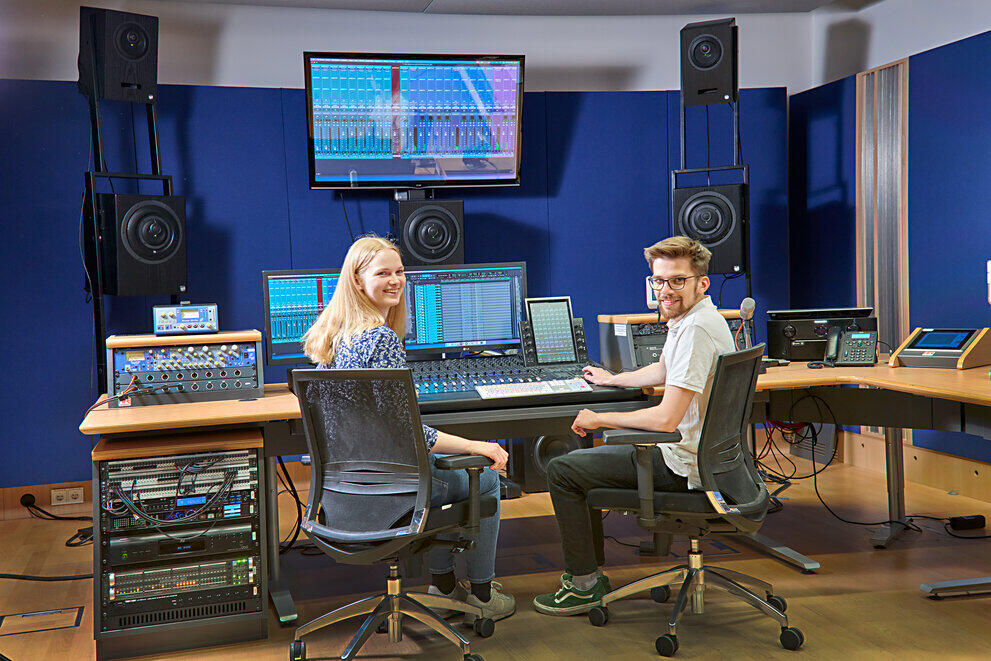 Zwei Studierende sitzen in einem Tonstudio mit viel Technik vor großen Bildschirmen und lächeln in die Kamera.