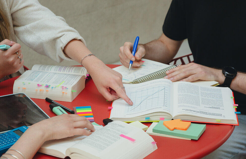 Three students are sitting at a table on which there are several textbooks, each marked with coloured sticky notes. One of the students is pointing to a diagram in one of the books.