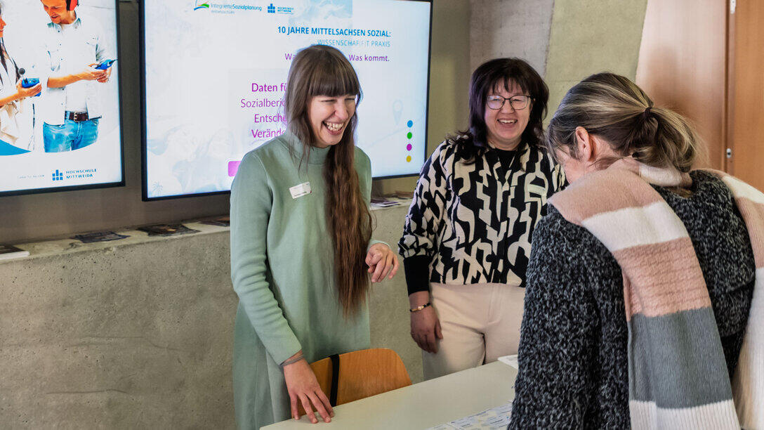 Two women, smiling warmly as they greet a participant. Behind them is a screen displaying the ‘Mittelsachsen Sozial’ logo.