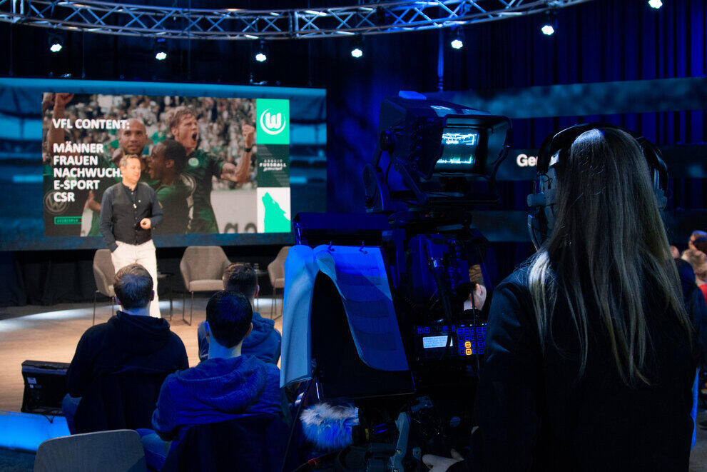 Eine Studentin steht hinter einer großen Fernsehkamera und filmt einen Mann auf der Bühne eines TV-Studios.