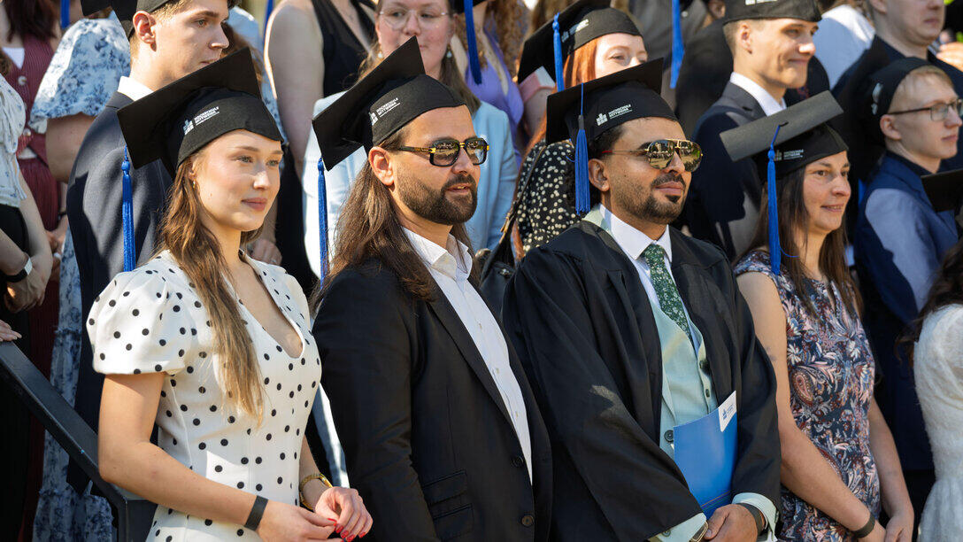 Vier Absolvent:innen mit Graduiertenhut stehen in einer Reihe, hinter ihnen weitere