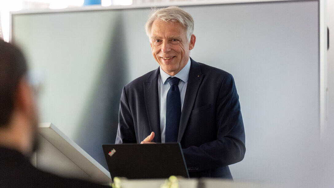 A man in a suit is standing in front of a screen, speaking with a smile. In the foreground of the picture, the head of a person seen from behind is visible, out of focus, in a half-length shot.