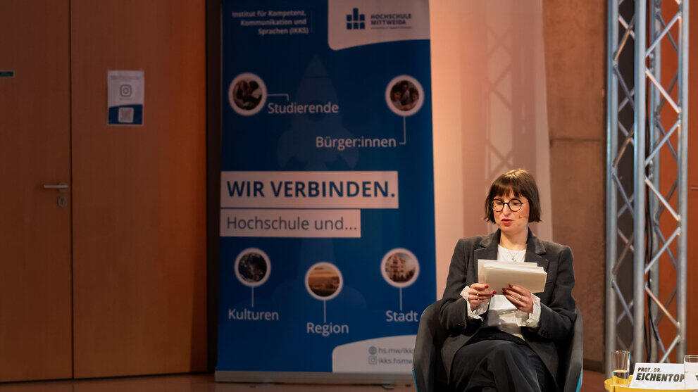 Weibliche Person sitzend mit Moderationskarten. Im Hintergrund ein blaues Rollup unter anderem mit dem Text "Wir verbinden Hochschule und ... ".