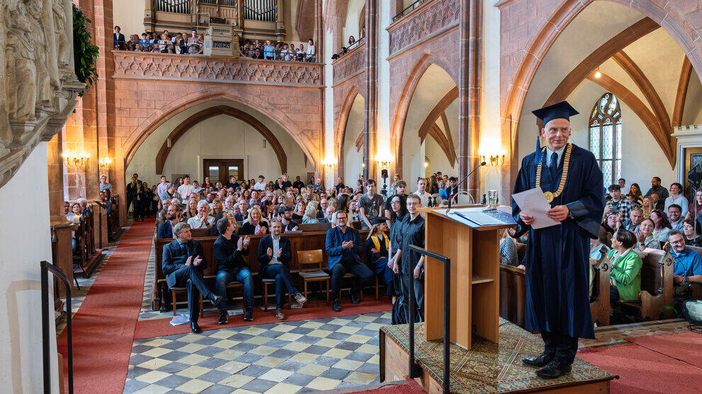 Blick in das vollbesetzte Kirchenschiff. Am Rednerpult stehend wendet sich eine männliche Person im blauen Talar, mit Doktorhut und goldener Amtskette den hinter im sitzenden Personen zu, die aber nicht im Bild zu sehen sind.