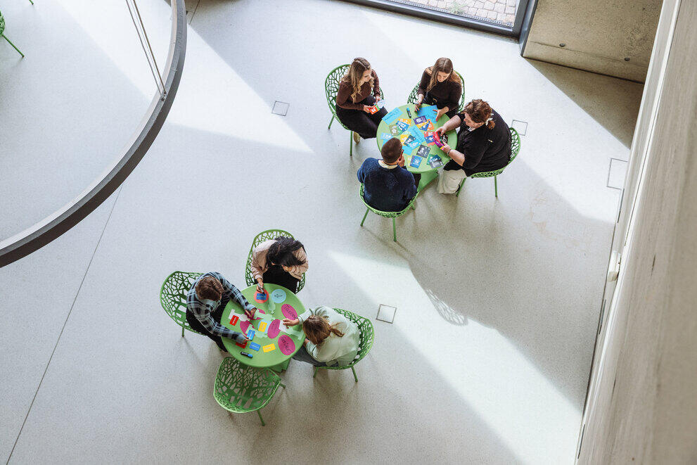 Sieben Studierende sitzen an zwei grünen Tischen und führen Gruppenarbeiten mit bunten Karten durch.