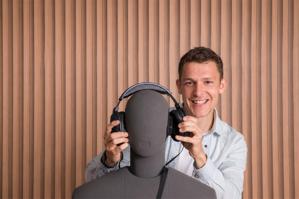 Ein lächelnder Student setzt einem Dummy Over-Ear-Kopfhörer auf.