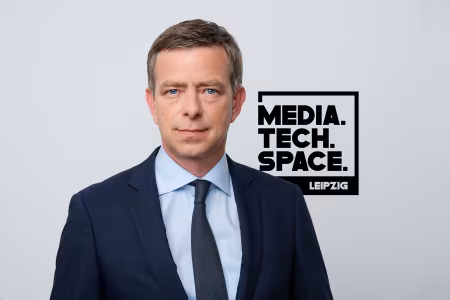 Ein Mann im Anzug mit Hemd und Krawatte guckt in die Kamera. Rechts ist das Logo des MediaTechSpace sichtbar.