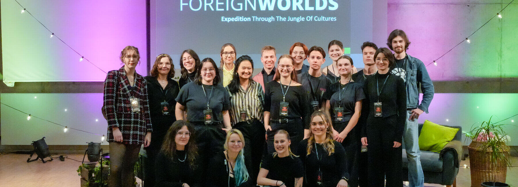 Gruppenbild von 19 jungen Personen in einer dekorierten Studioatmosphäre, von den einige knien. Im Hintergrund ist auf einer Leinwand der Schriftzug „Foreign Worlds. Expedition Through The Jungle of Cultures“ zu lesen.