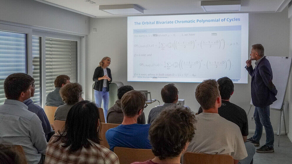Eine weibliche Dotenten links und ein männlicher Dozent rechts von einer projizieren Folie mit einer mathematischen Tabelle. Im Vordergrund sind Zuhörende von hinten zu sehen.