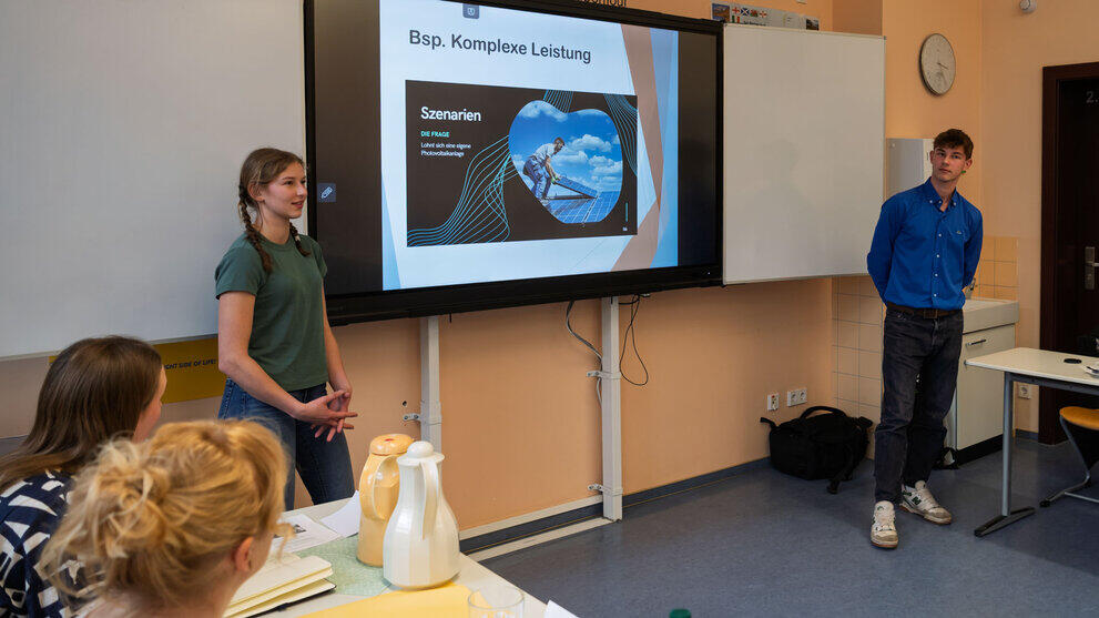 Schülerin und Schüler stehen llinks und rechts von einem Screen mit einer Folie mit der Überschrift "Beispiel Komplex Lesitung".