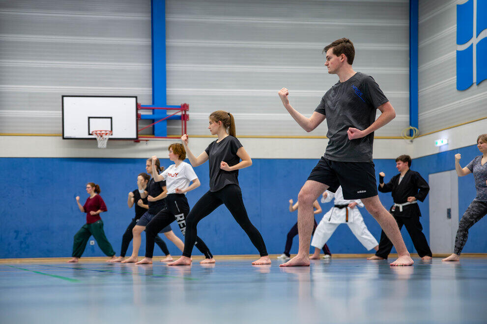 Circa zehn Student:innen machen gemeinsam Karate. Sie führen Zenkutsu-dachi mit Uchi-Uke aus, eine Abwehrtechnik gegen Angriffe.