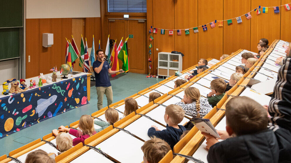 Moderator steht vor Publikum in Hörsaal mit internationalen Flaggen und Vernetzung auf Leinwand.