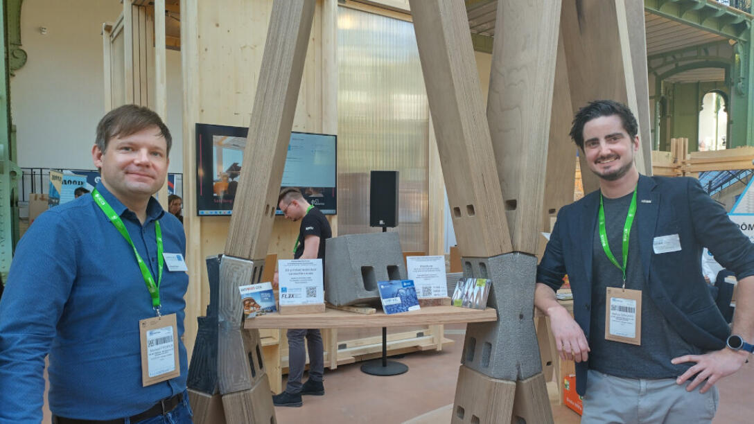 zwei männliche Personen stehen an einer Holzkonstruktion und lächeln in die Kamera