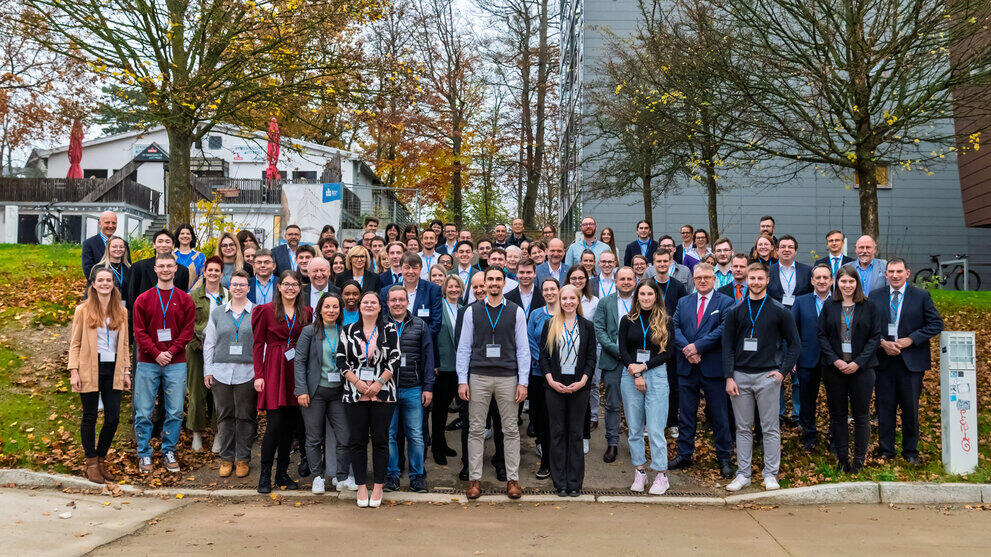 Eine Menschengruppe mit rund 90 überwiegend lachenden Personen steht auf einem ansteigenden Weg und dem Rasen links und rechts davon. Im Hintergrund befinden sich Gebäude und weitgehend entlaubte Bäume.