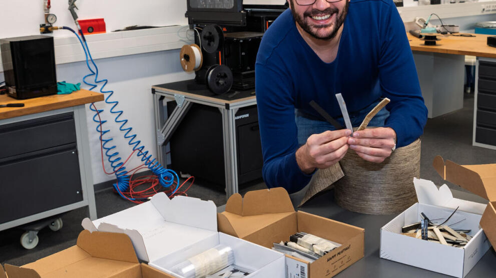 A man in a laboratory is smiling cheerfully as he holds up two strip-shaped material samples towards the camera. On the table in front of him are boxes containing further samples.