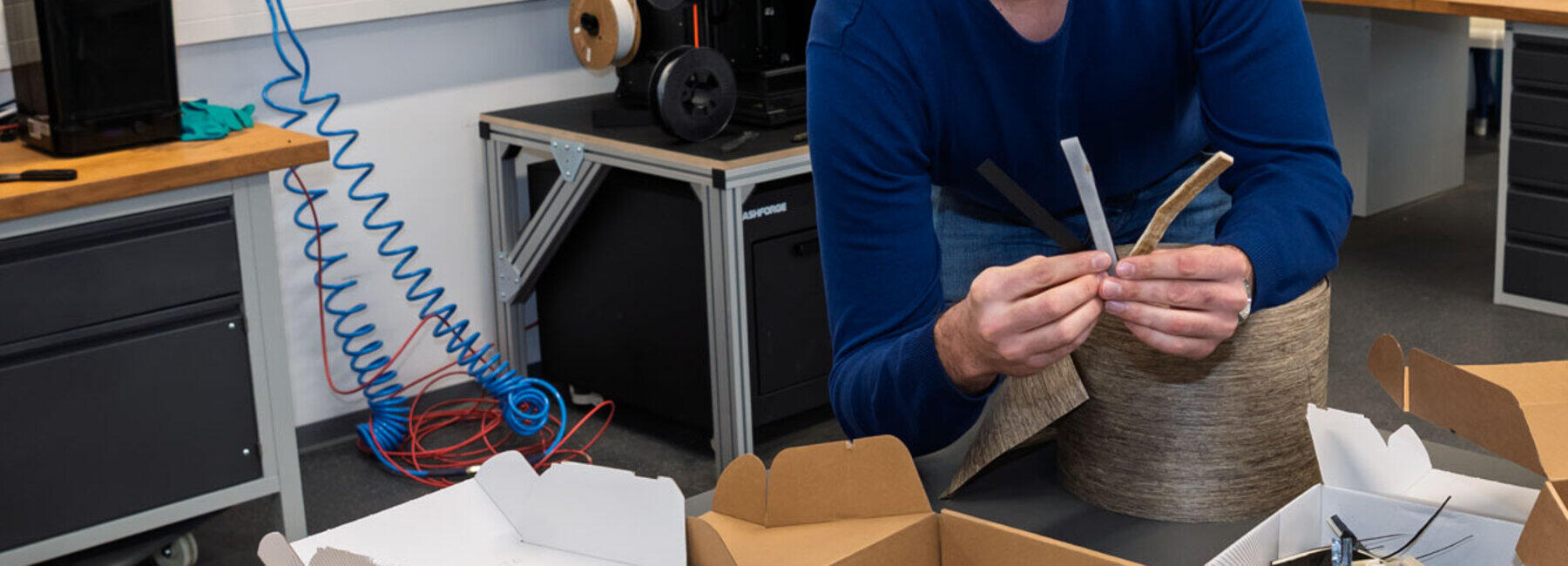 A man in a laboratory is smiling cheerfully as he holds up two strip-shaped material samples towards the camera. On the table in front of him are boxes containing further samples.