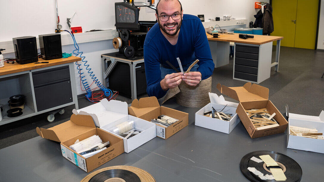 Tisch mit Bauteilen, männliche Person beugt sich darüber und schaut lächelnd in die Kamera