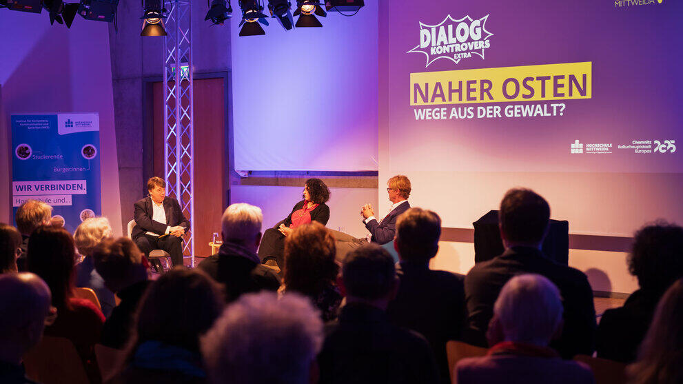 Blicl über die Köpfe von Zuschauenden hinweg auf drei Personen des Podiusm. Über ihnen auf der Leinwand ist "Dialog Kontrovers Extra. Naher Osten. Wege aus der Gewalt" zu lesen".